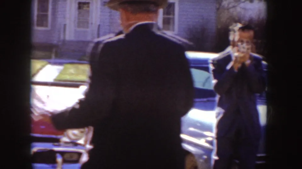1955 Idaho street scene with a hatted suited man walking past and another man using a camera by parked cars.