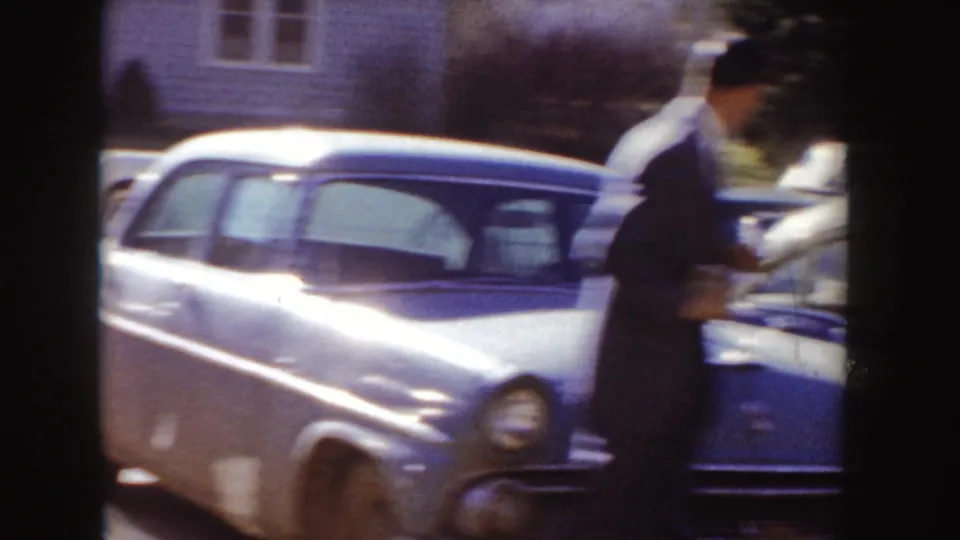 1955 Idaho home-movie frame showing a suited man beside a light-colored classic car with houses behind.