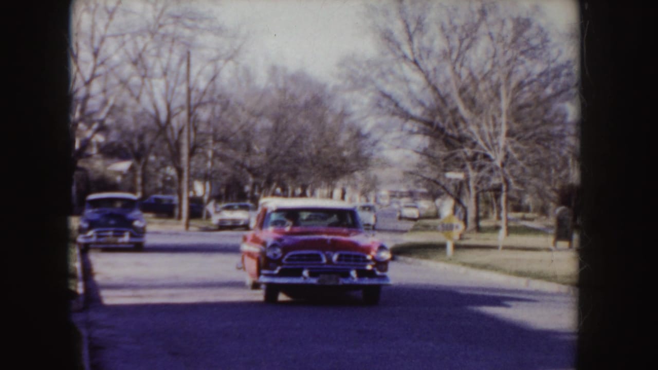 Red mid-century sedan approaches on a quiet residential street with parked cars and bare trees.