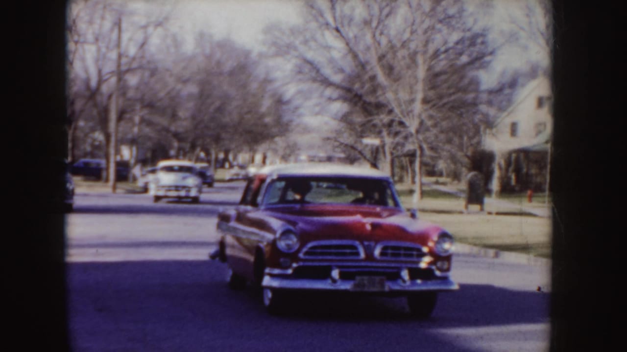 Red sedan nears the camera with another car behind it on a leafless suburban block.