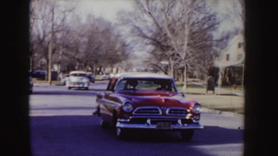 The red classic sedan draws closer on an Idaho residential road, with another period car and bare street trees visible behind it.