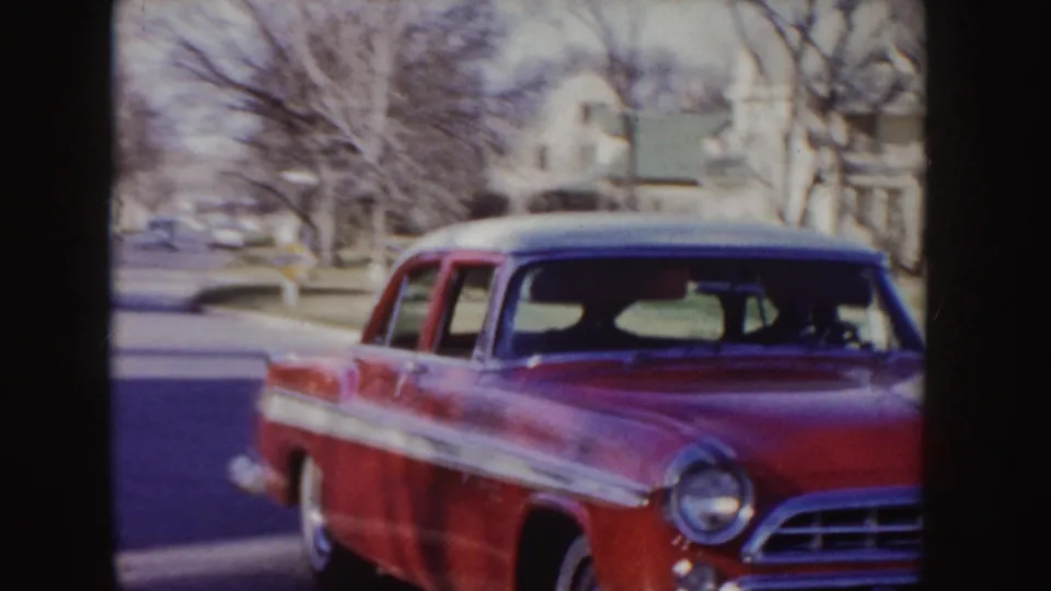 Close 1955 Idaho home-movie view of the red sedan as it passes near the camera, with houses, trees, and roadway behind it.