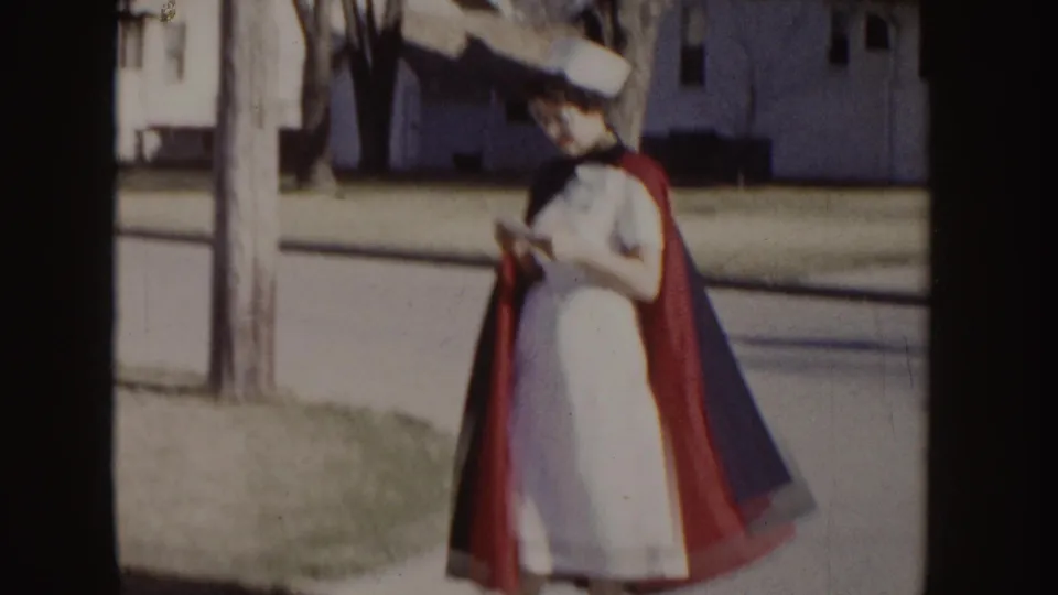 1955 Idaho home-movie frame of a woman in a white nurse uniform, cap, and red cape standing along a suburban curb.