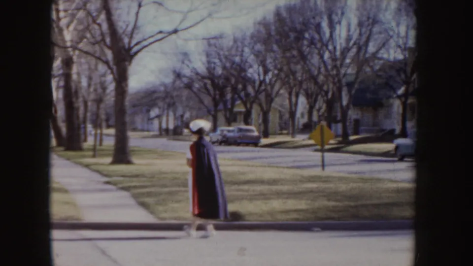 Wide 1955 Idaho frame showing the uniformed woman in a red cape near a curb on a residential street with parked cars and leafless trees.