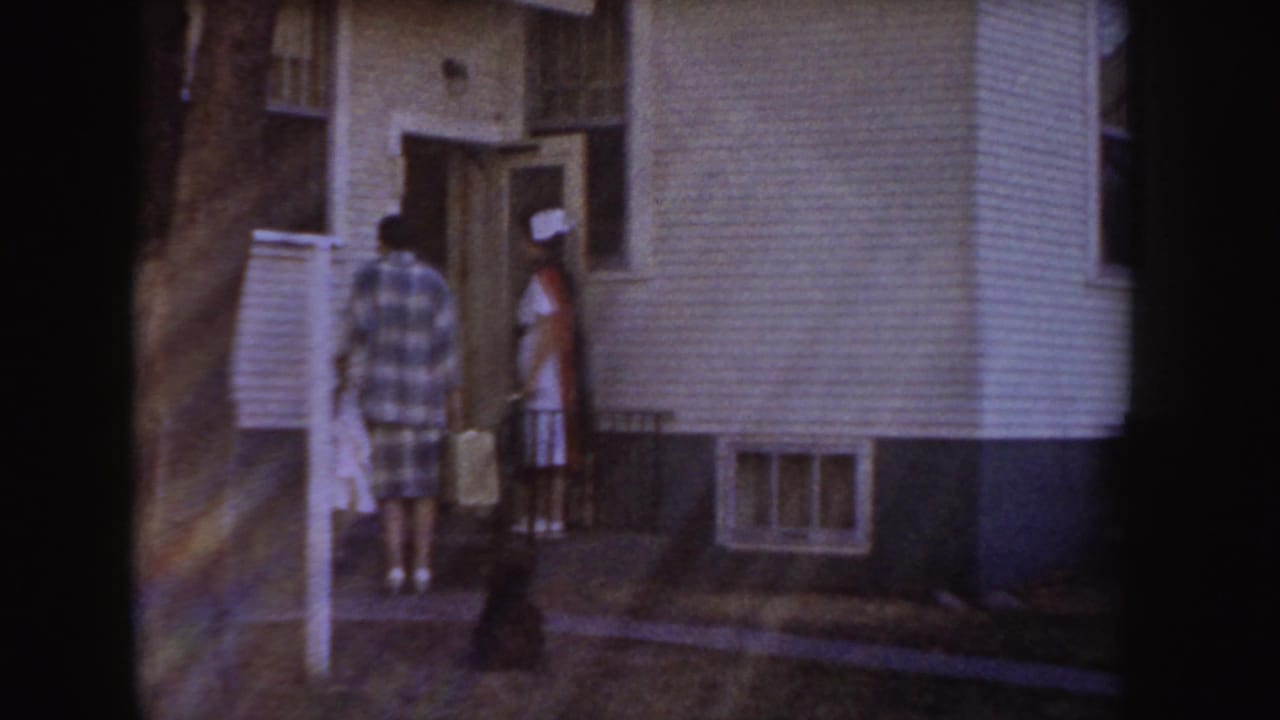 Two people stand near the open doorway of a white-sided house.