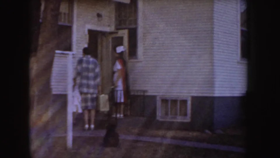 In 1955 Idaho home-movie footage, a man and a woman in a white cap stand outside a modest white-sided house doorway.