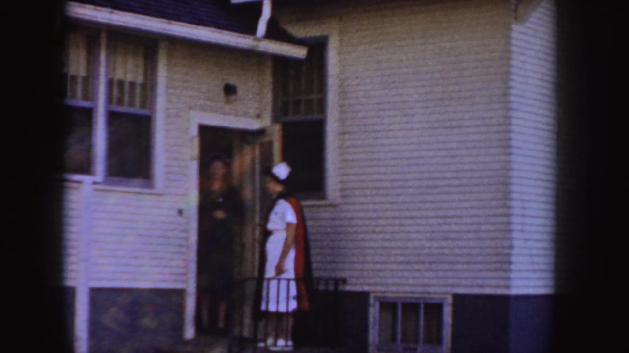 A woman in a white cap stands at an open house door with another person inside.