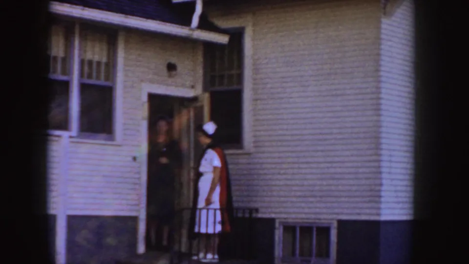 A woman in a nurse-style cap stands at the doorway of a white-sided Idaho house, with another person visible just inside.