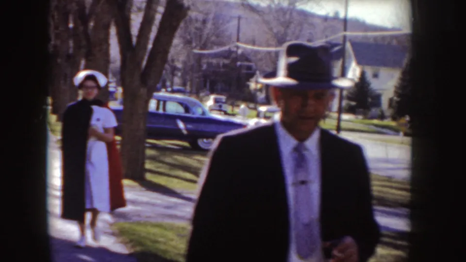 A man in a dark suit and fedora passes the camera while a woman in a white cap stands near the sidewalk in a 1955 Idaho neighborhood.