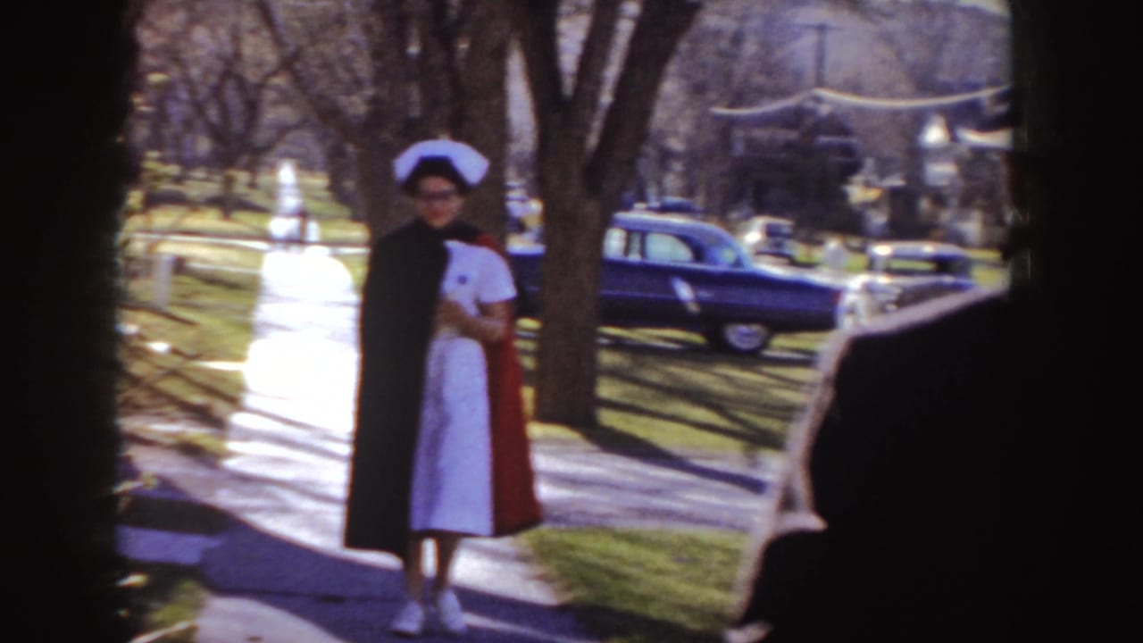 Nurse in a white uniform stands on a sidewalk near a red station wagon and bare trees.