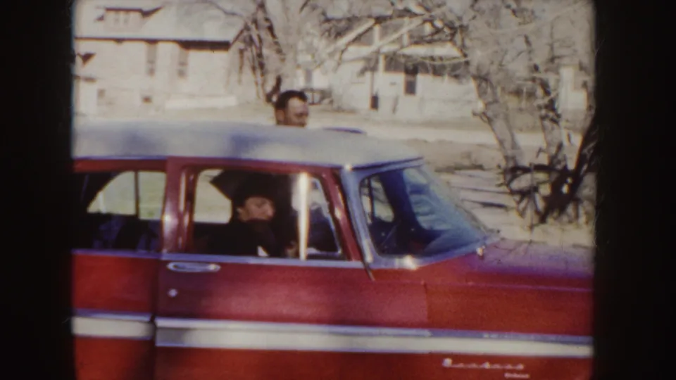 A red station wagon with white trim is parked on a residential street, with one man visible inside and another standing behind the car.