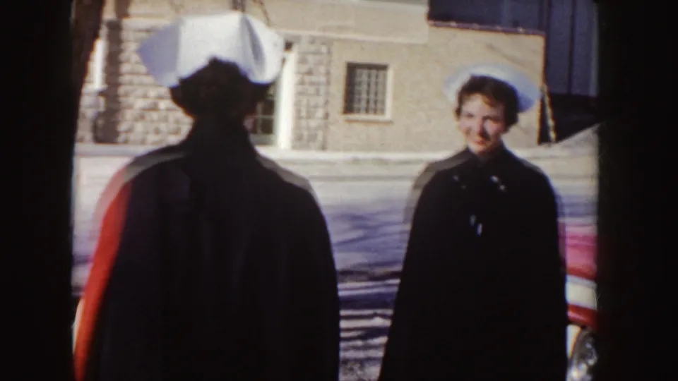 A nurse in a white cap and cape-like coat faces a smiling man on a residential street beside the red station wagon.