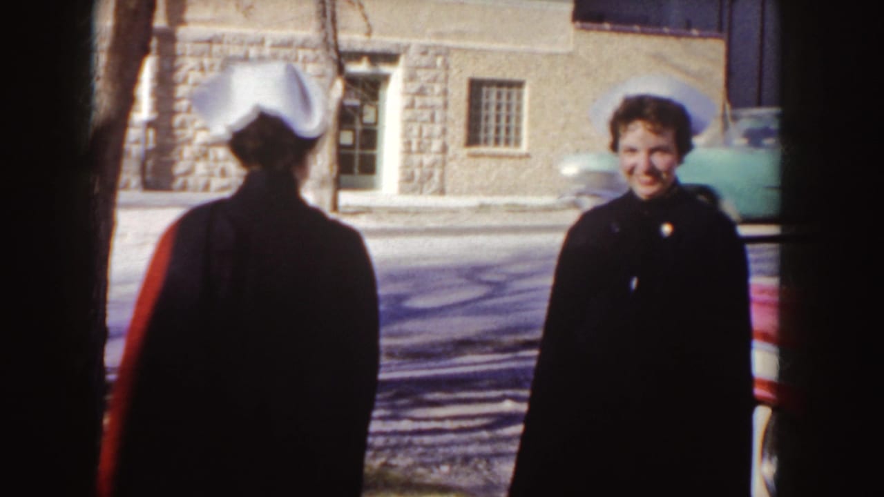 Two women in white nurse caps stand near a red car on a town sidewalk.