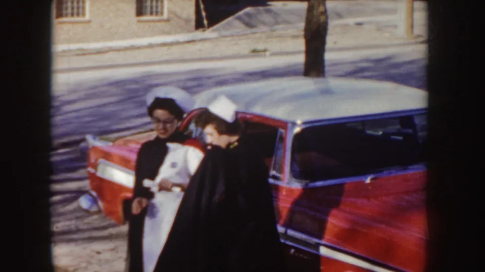 Two women in white nurse caps smile beside a red and white 1950s sedan in an Idaho town setting.
