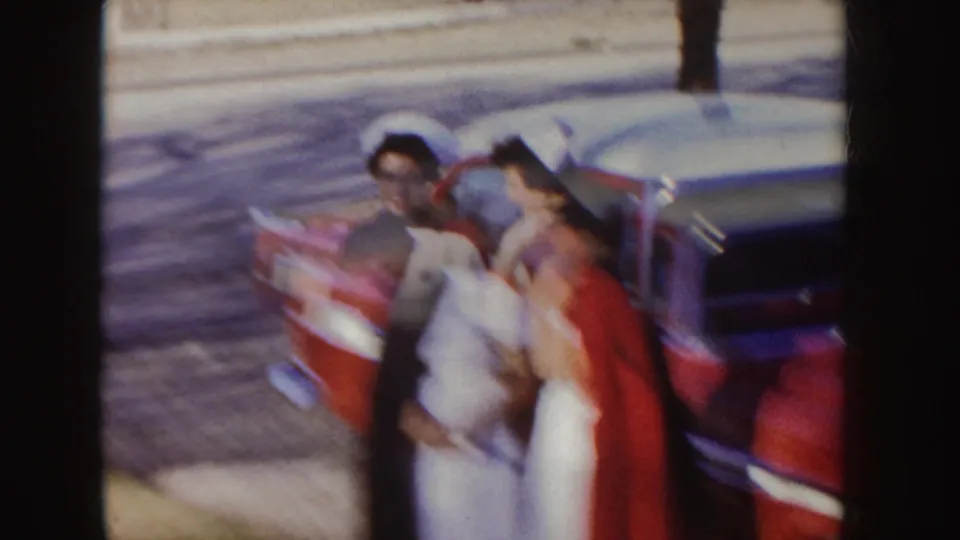 Soft 8mm frame of two women in nurse caps moving close to a red sedan during a filmed town-sidewalk moment.