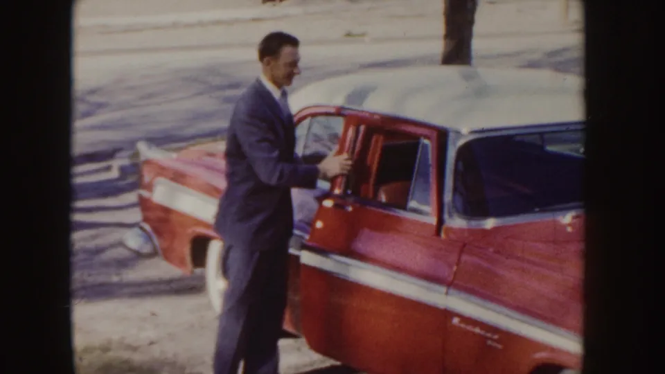 A suited man opens the door of a red and white 1950s sedan on a small-town Idaho street.
