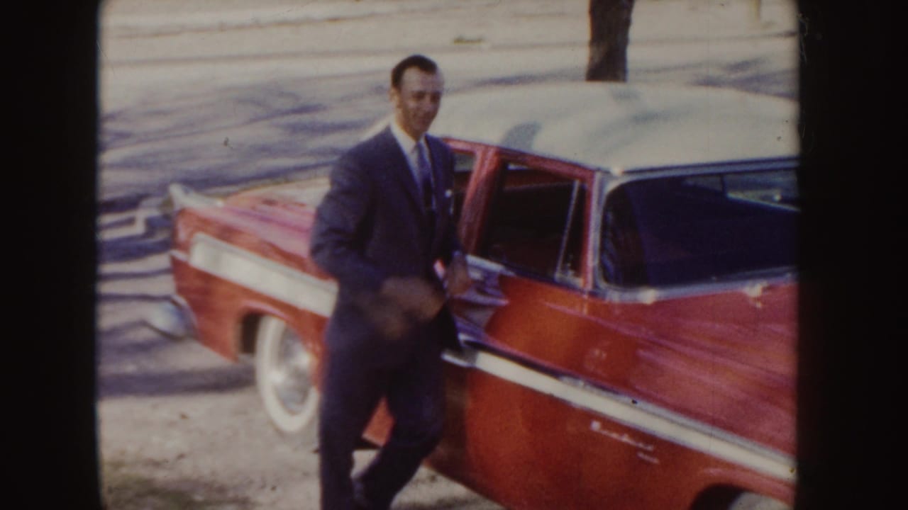 Man in a dark suit standing beside a red and white 1950s car with the driver door open.