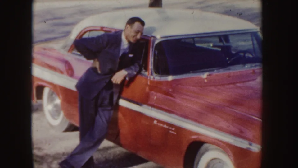 A man in a dark suit leans into the open driver door of a red and white 1950s car in the Idaho home-movie scene.