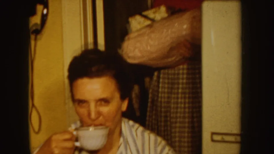 A woman in a striped shirt sits indoors and raises a teacup in warm 1955 Idaho home-movie footage.