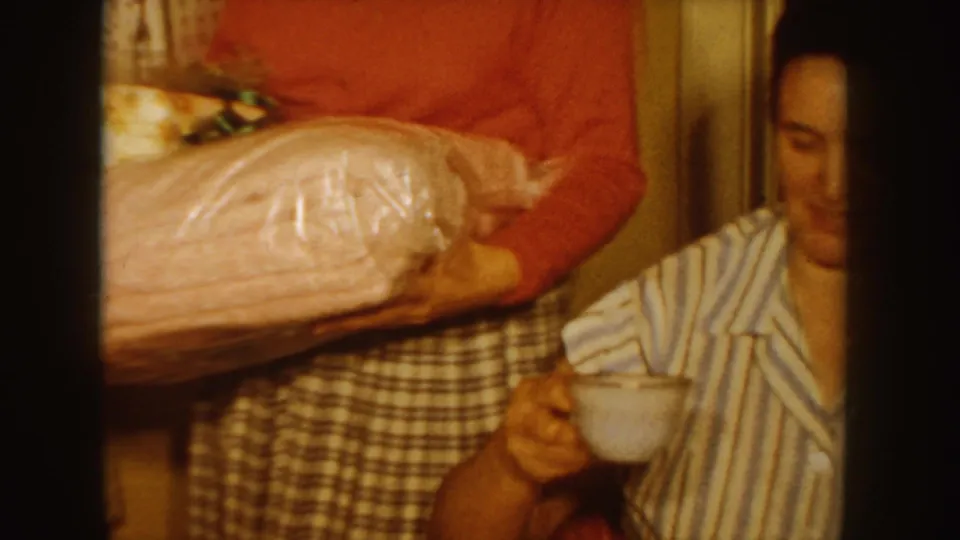 1955 Idaho home-movie frame showing a woman in a red sweater carrying wrapped presents toward a seated girl in striped pajamas.