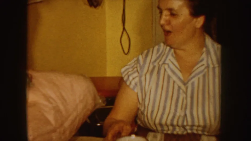 1955 Idaho frame of a seated girl in striped pajamas reacting beside bedding or cushions in a warm private interior.