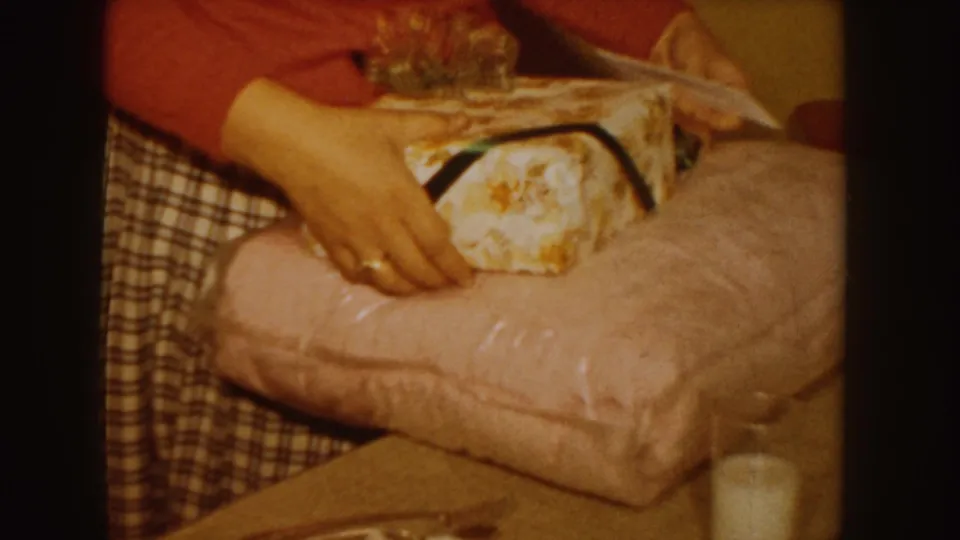 1955 Idaho home-movie frame showing hands placing a wrapped present on pink bedding, with a glass of milk nearby.