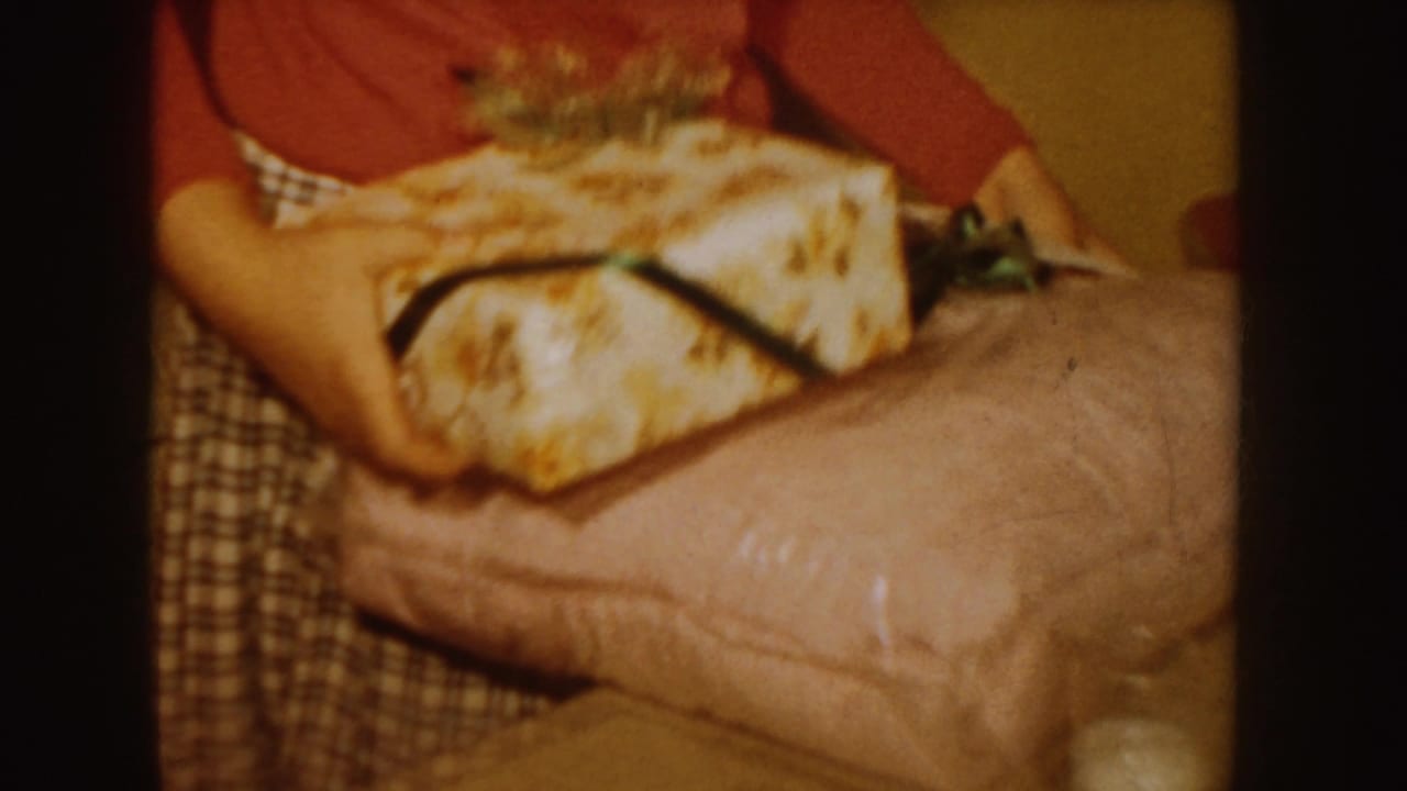 In a 1955 Idaho home-movie frame, an adult woman in a red sweater and plaid skirt handles a wrapped gift with ribbon indoors.