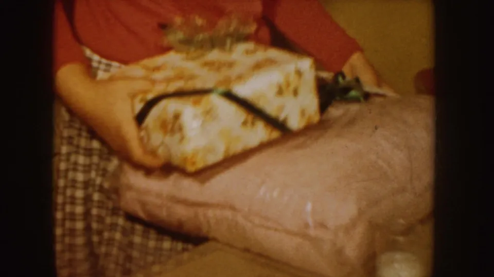 In a 1955 Idaho home-movie frame, an adult woman in a red sweater and plaid skirt handles a wrapped gift with ribbon indoors.
