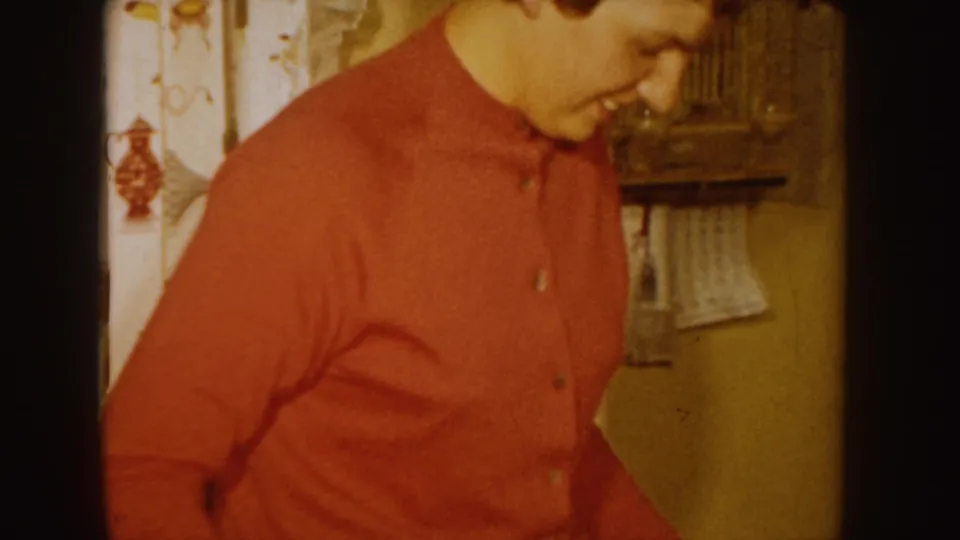 An adult woman in a red sweater looks downward in a private Idaho interior, with patterned curtains and household furnishings behind her.