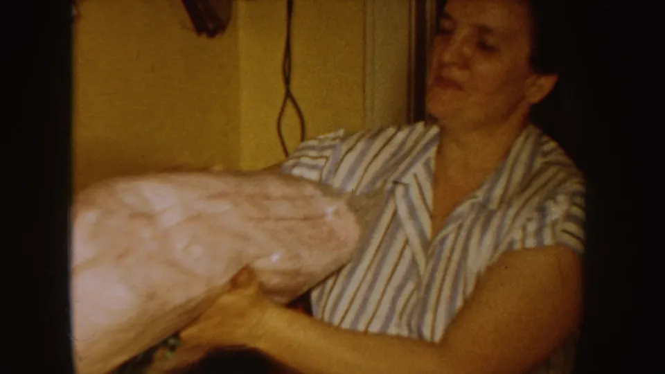 An adult woman in striped pajamas holds a large pink pillow in a private home interior, seen in warm faded 8mm color.