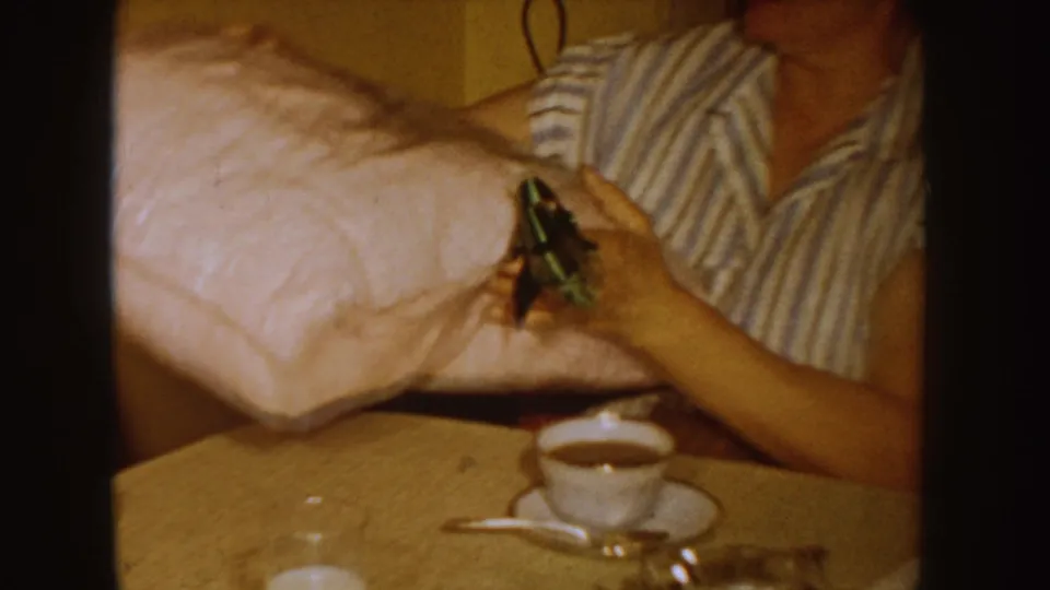 A woman in striped pajamas holds a pink pillow beside a table with a teacup and saucer in the 1955 Idaho home-movie scene.