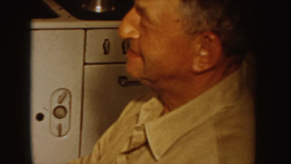In a 1955 Idaho home-movie frame, a seated man in a collared shirt is seen in profile near mid-century kitchen cabinetry.