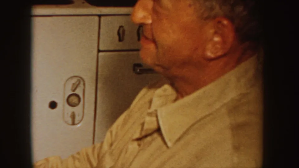 A 1955 Idaho home-movie frame shows a close profile view of a seated man in a collared shirt beside kitchen cabinets.