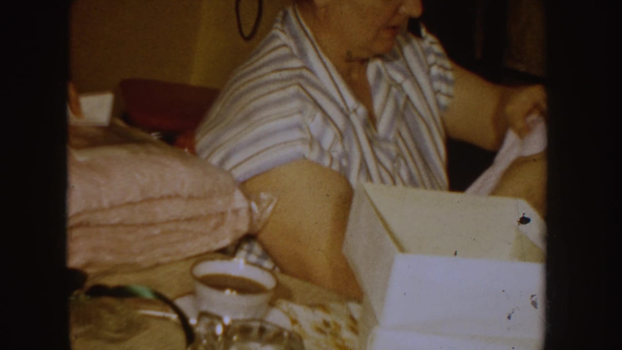 In a 1955 Idaho home-movie frame, a woman in a striped shirt folds fabric near a bed with a coffee cup on a nearby surface.
