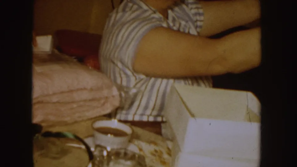 In this 1955 Idaho home-movie frame, a woman in a striped shirt reaches forward in a bedroom-like interior with bedding and a coffee cup visible.