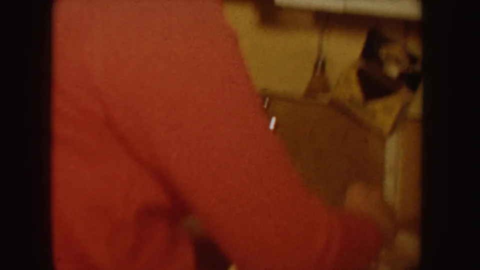 A red-sweatered figure appears in soft focus inside the 1955 Idaho home-movie interior, with bedding and household objects partly visible.
