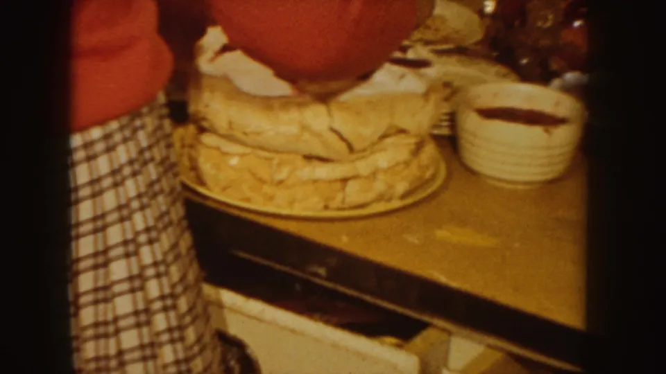 In a 1955 Idaho home-movie frame, a woman in red and a checkered apron works over a layered cake with white frosting on a table.