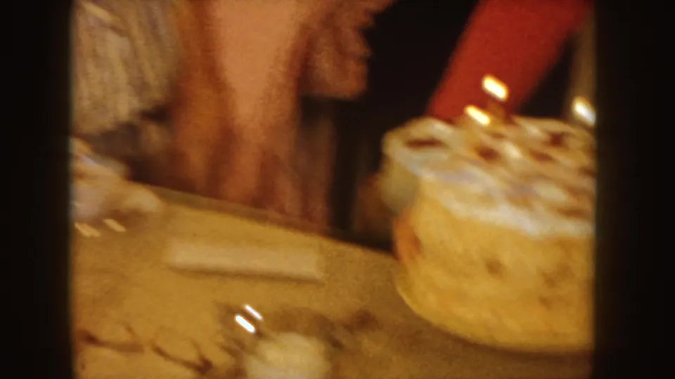 A white frosted cake with lit candles is placed near the edge of a tan table in a 1955 Idaho home interior.