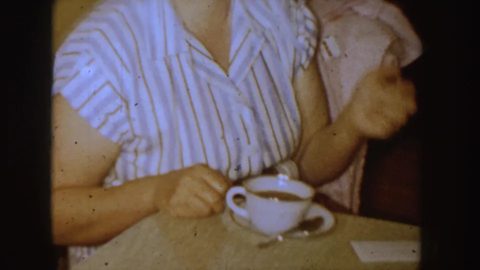 A woman in a striped house dress holds a white teacup at a wooden dining table in this 1955 Idaho home-movie frame.