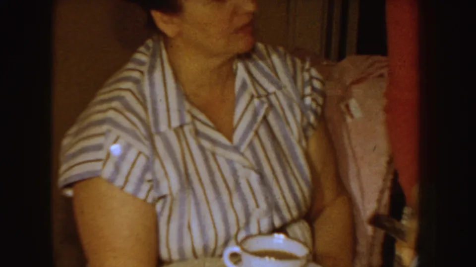 The woman sits beside a teacup at the dining table, partly turned away, in warm faded 1955 Idaho 8mm footage.
