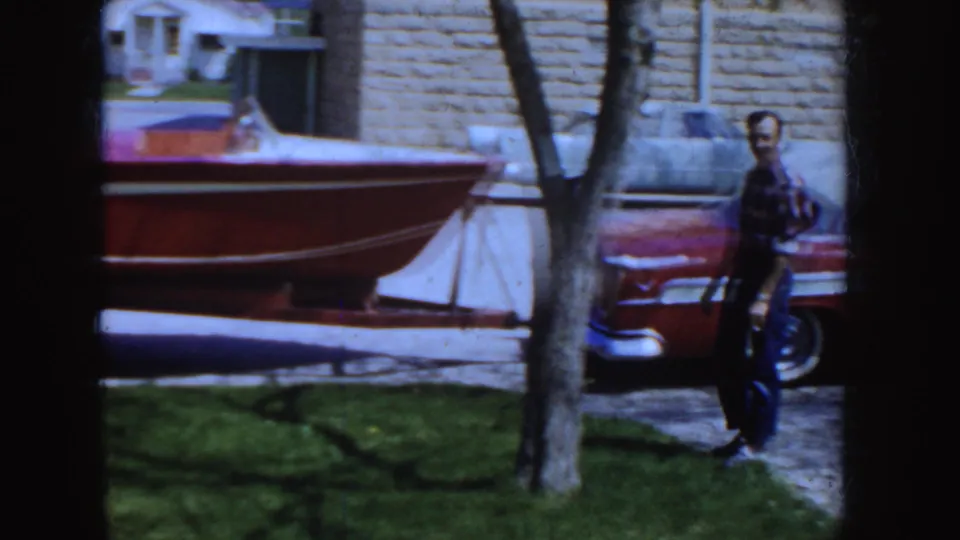 1955 Idaho home-movie frame with a man beside a red-and-white sedan and small boat on a trailer near a driveway.