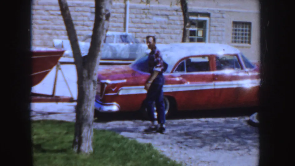 1955 Idaho 8mm frame showing a man walking in a driveway beside a red-and-white sedan and nearby boat trailer.