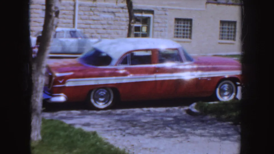 1955 Idaho home-movie frame centered on a red-and-white mid-century sedan parked outside near a tree and building wall.