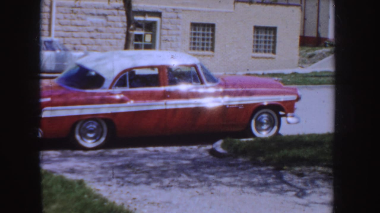 Red-and-white mid-1950s sedan parked on an Idaho residential street, with a masonry house and driveway visible behind it.