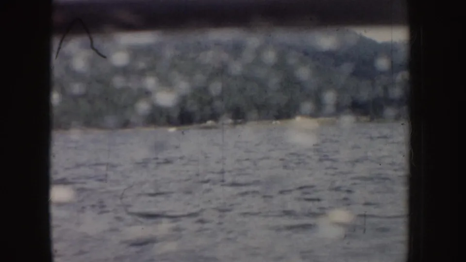 1955 Idaho 8mm frame with rippled lake water, a distant wooded mountain slope, pale sky, and dark rail divisions across the view.