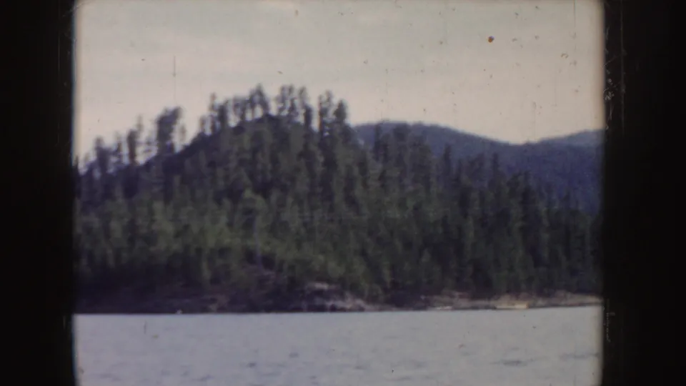 1955 Idaho home-movie view of a green wooded island across rippled lake water, with distant hills under a pale sky.