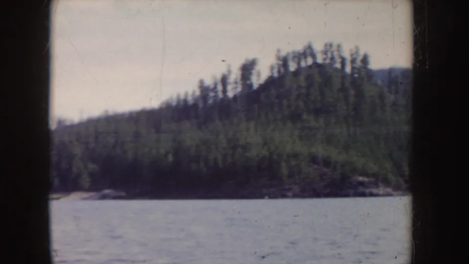 1955 Idaho 8mm still of a tree-covered island shoreline sliding past above a rippled lake surface.
