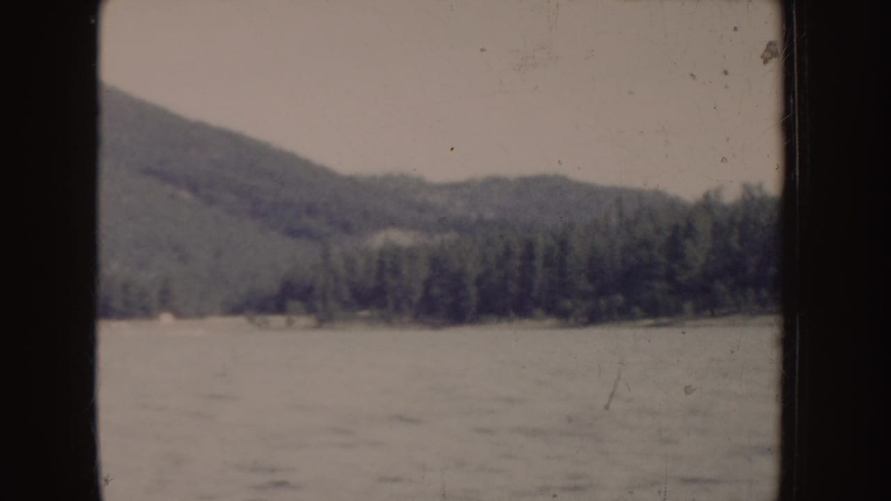 Forested hillside and shoreline seen across rippling water with dark film borders.