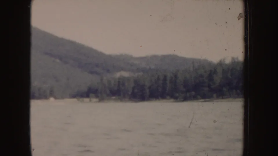 1955 Idaho 8mm frame showing a tree-covered hill and shoreline across open water, viewed from a boat.