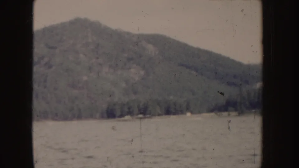 1955 Idaho home-movie frame showing a forested slope and lakeshore from a moving boat perspective.